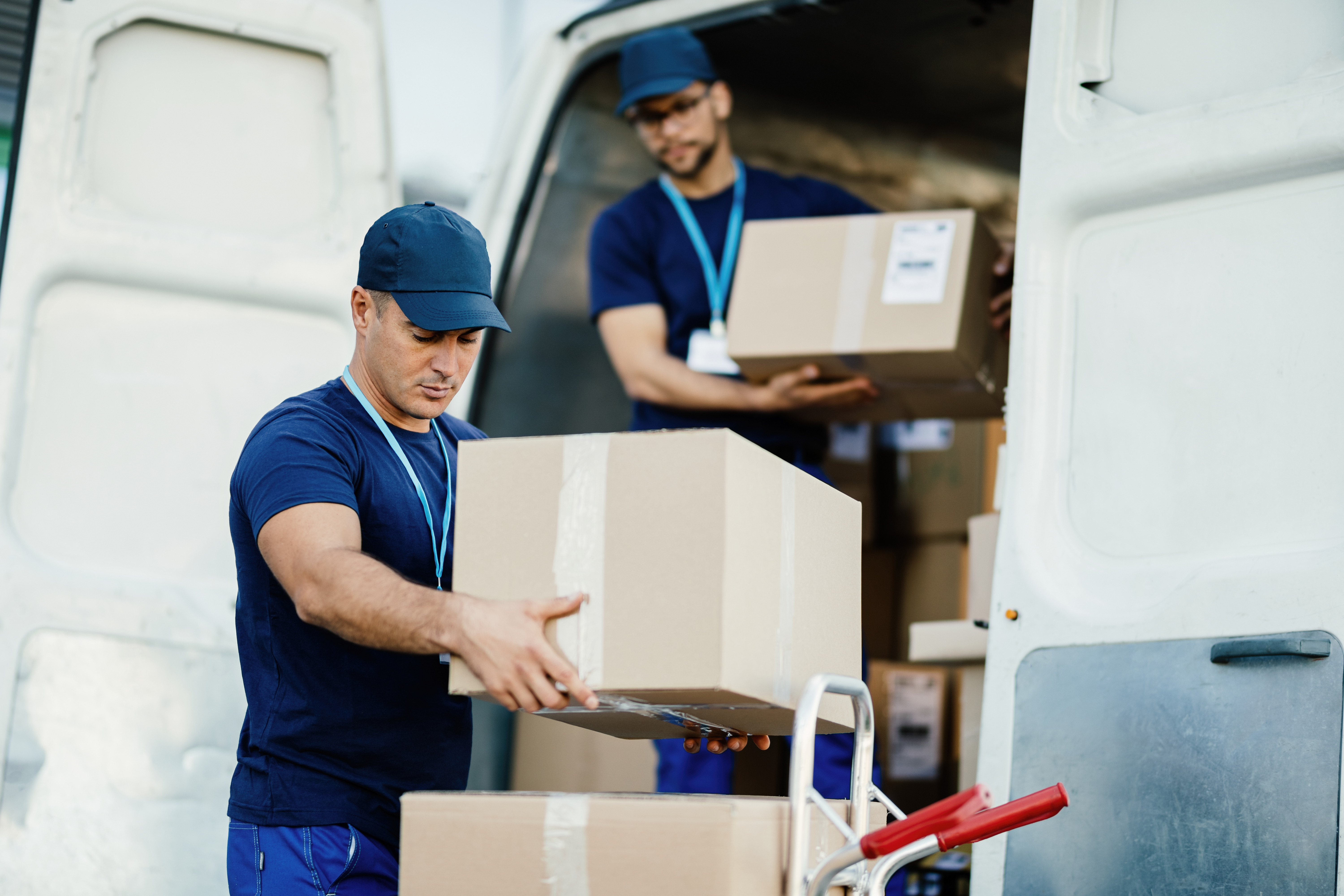 young courier and his colleague unloading cardboard boxes from delivery van.